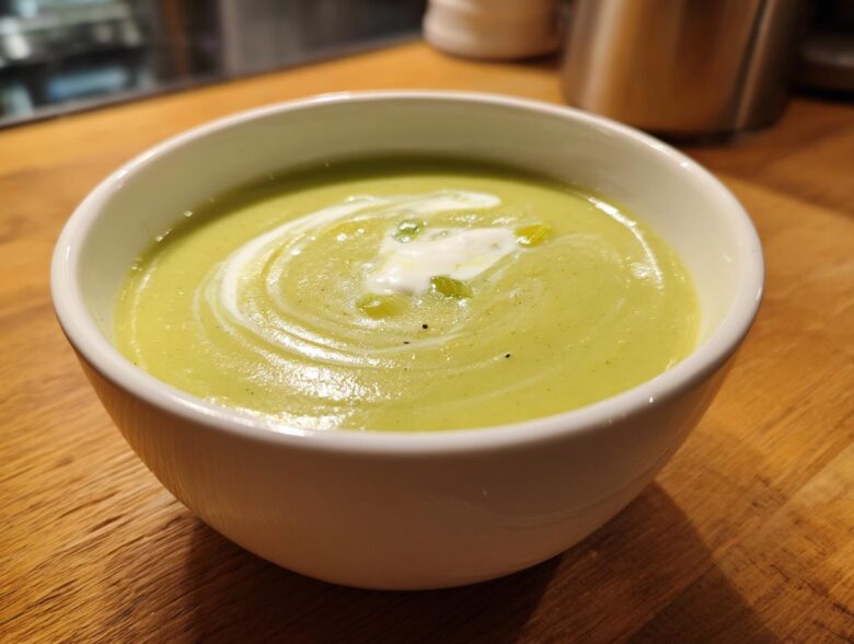 A close-up of a white bowl filled with smooth, light green leek soup, garnished with a swirl of cream and olive oil.