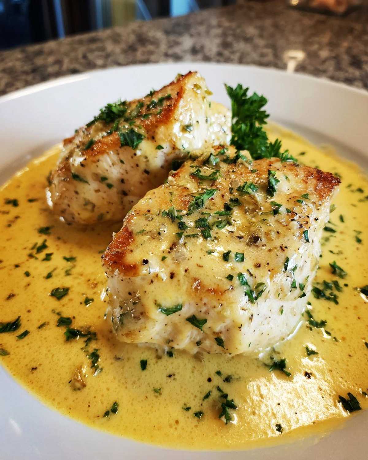 Two pieces of seared chicken breast smothered in a rich, yellow creamy lemon chicken sauce and topped with fresh parsley.
