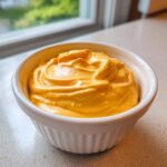 A close-up of thick, bright orange homemade nacho cheese sauce swirled in a small white ramekin.