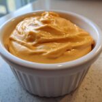 Close-up of rich, orange, creamy nacho cheese sauce filling a small white ribbed ramekin.