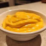 Close-up of rich, smooth, bright orange nacho cheese sauce served in a small white dipping bowl.