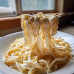 A fork lifts dripping, creamy fettuccine coated in rich Olive Garden Alfredo Sauce from a white bowl.
