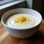 A close-up of creamy roasted cauliflower soup topped with bright yellow lemon zest in a white bowl.