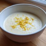 A close-up of creamy roasted cauliflower soup topped with bright yellow lemon zest in a white bowl.