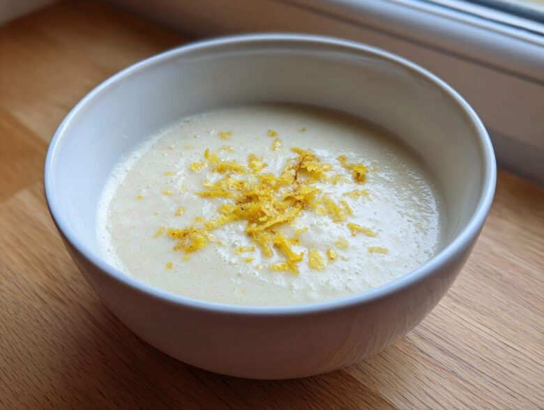 A close-up of creamy roasted cauliflower soup topped with bright yellow lemon zest in a white bowl.