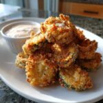A pile of golden brown, crispy Fried Pickles served on a white plate next to a small ramekin of creamy dipping sauce.