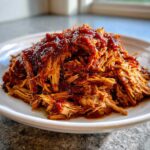 Close-up of tender, shredded crock pot pulled pork generously topped with rich, dark BBQ sauce on a white plate.