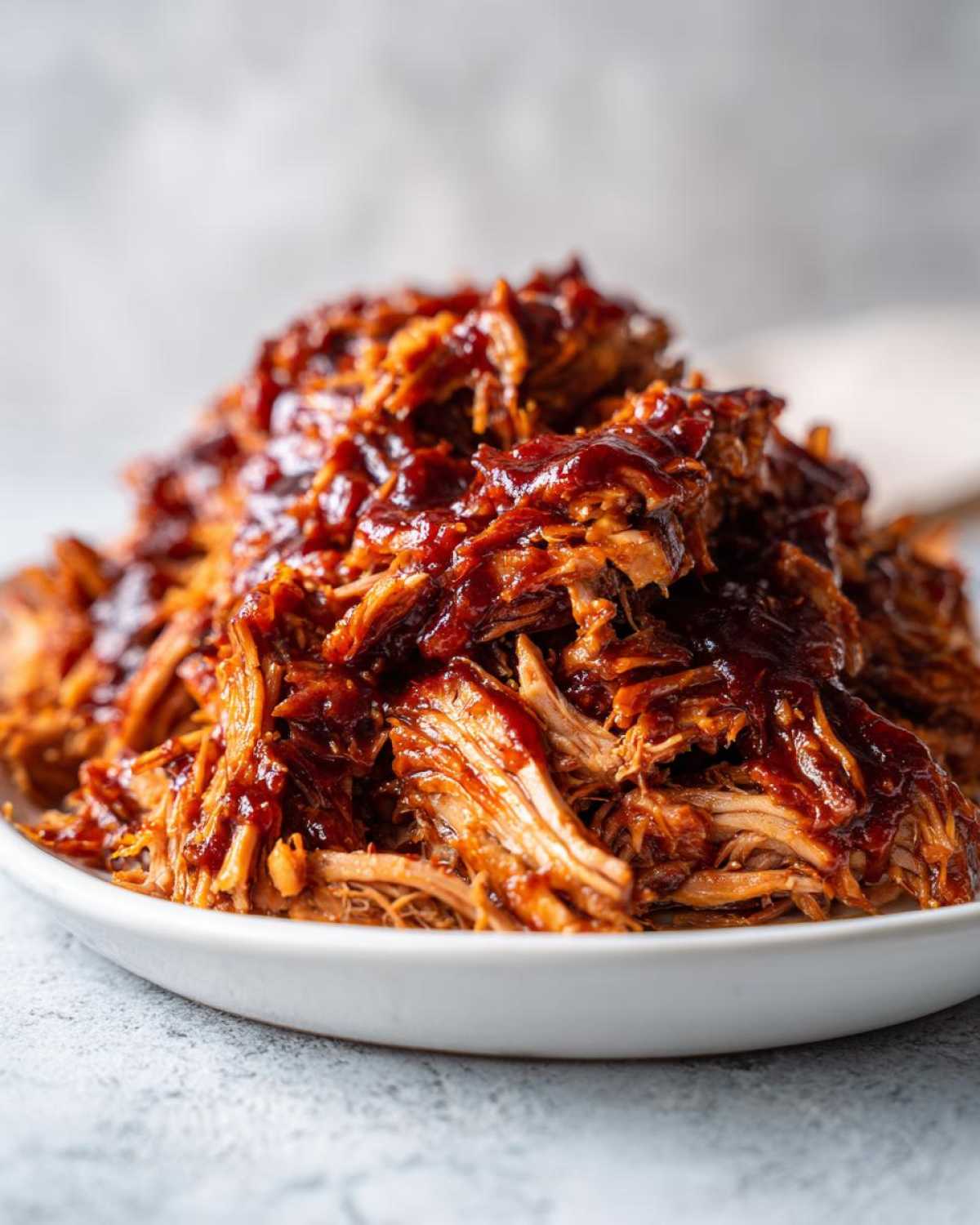Close-up of tender, shredded crock pot pulled pork generously coated in rich barbecue sauce on a white plate.