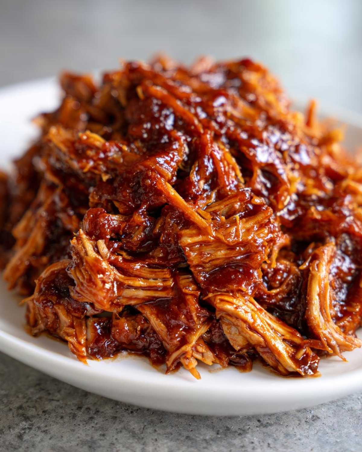 Close-up of tender, shredded crock pot pulled pork coated in a rich, glossy BBQ sauce served on a white plate.