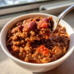 A spoonful of thick, meaty crockpot chili with visible kidney beans and tomato chunks being lifted from a white bowl.