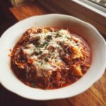 A close-up of a white bowl filled with rich crockpot lasagna soup, topped generously with melted mozzarella and fresh parsley.