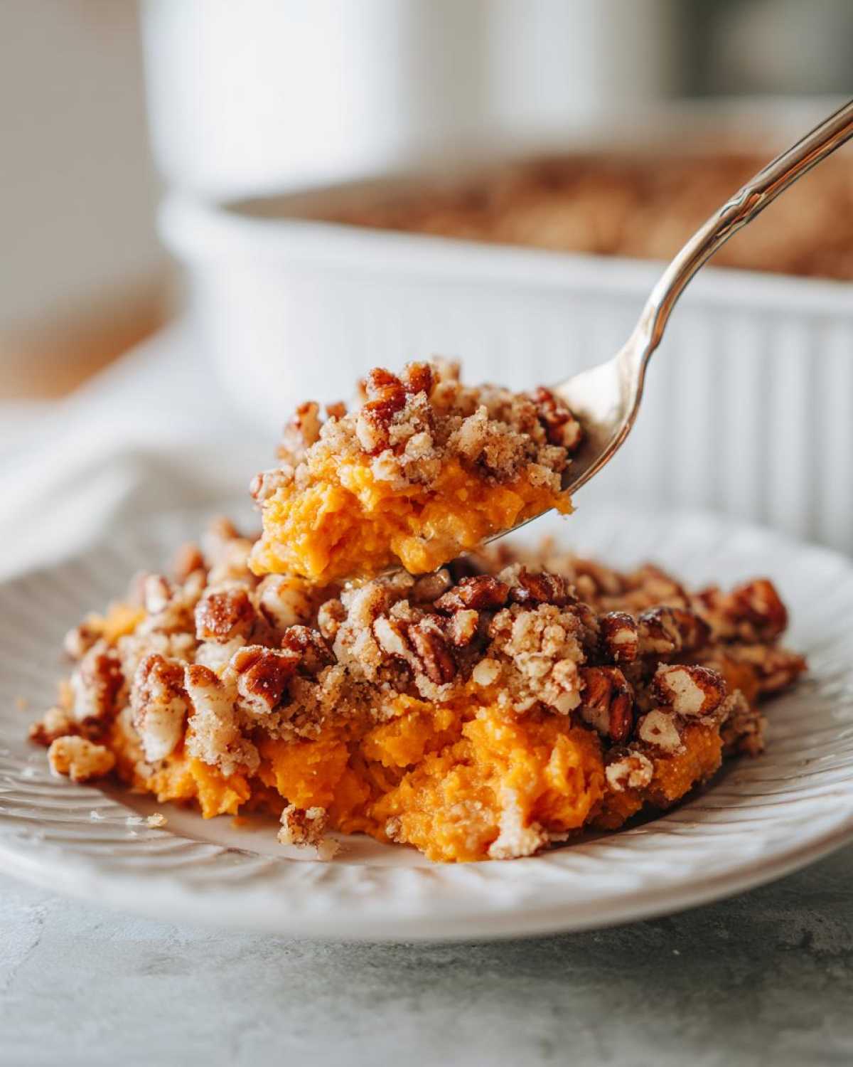 A spoonful of orange crockpot sweet potato casserole topped with crunchy pecans being lifted from a serving on a white plate.