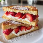 Two halves of decadent stuffed french toast stacked, filled with cream cheese and fresh sliced strawberries, dusted with powdered sugar.