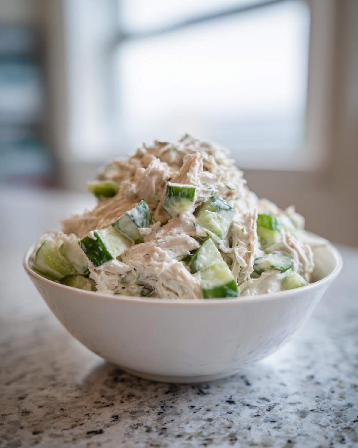 A white bowl filled high with creamy dill pickle chicken salad mixed with chunks of fresh cucumber.