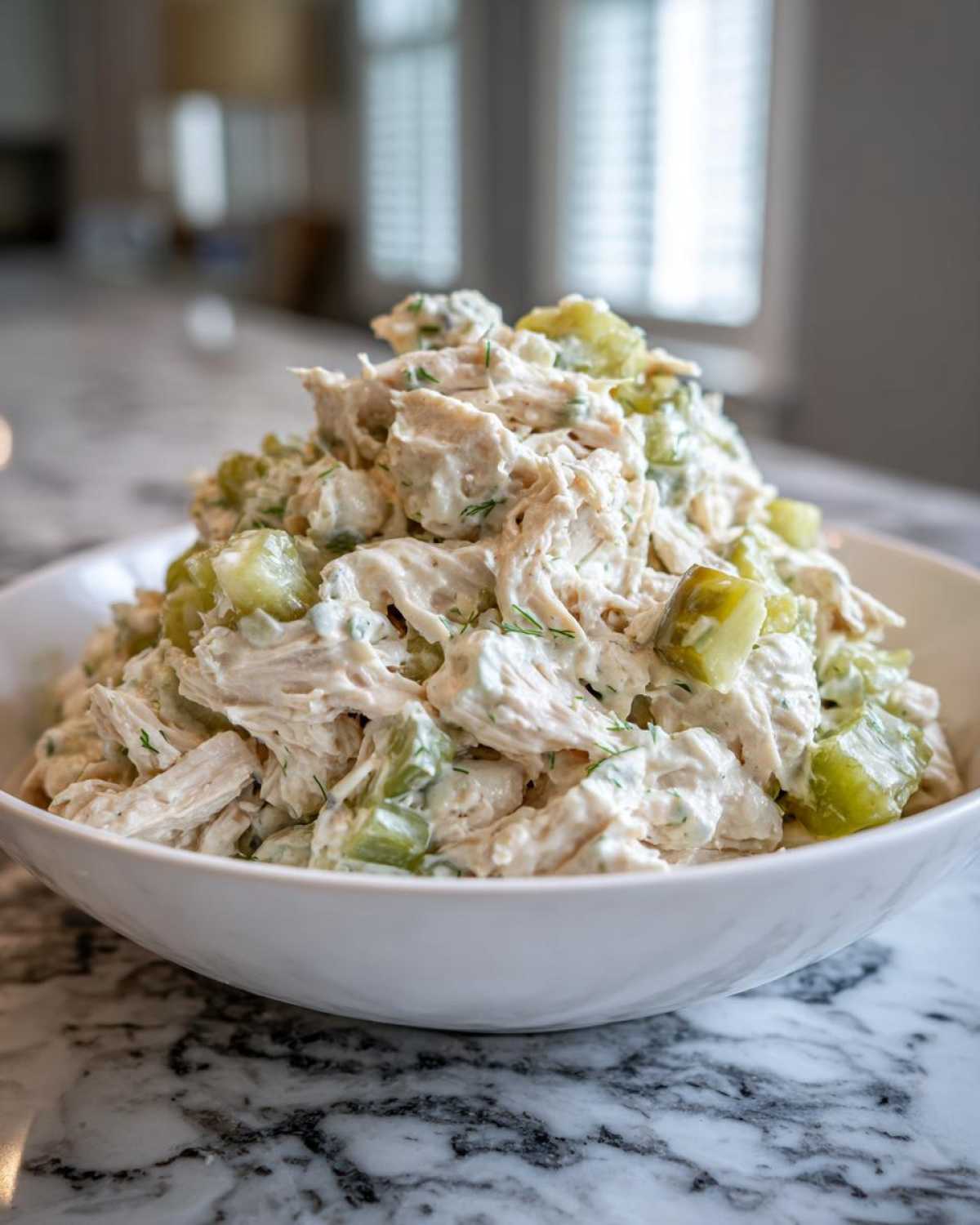 A generous serving of creamy dill pickle chicken salad piled high in a white bowl on a marble counter.