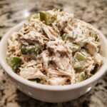 A close-up of creamy dill pickle chicken salad featuring shredded chicken and chunks of pickles in a white bowl.