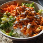Close-up of a vibrant buffalo chicken bowl featuring shredded chicken in orange sauce over rice, topped with ranch and shredded carrots.