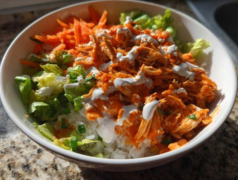 Close-up of a vibrant buffalo chicken bowl featuring shredded chicken in orange sauce over rice, topped with ranch and shredded carrots.