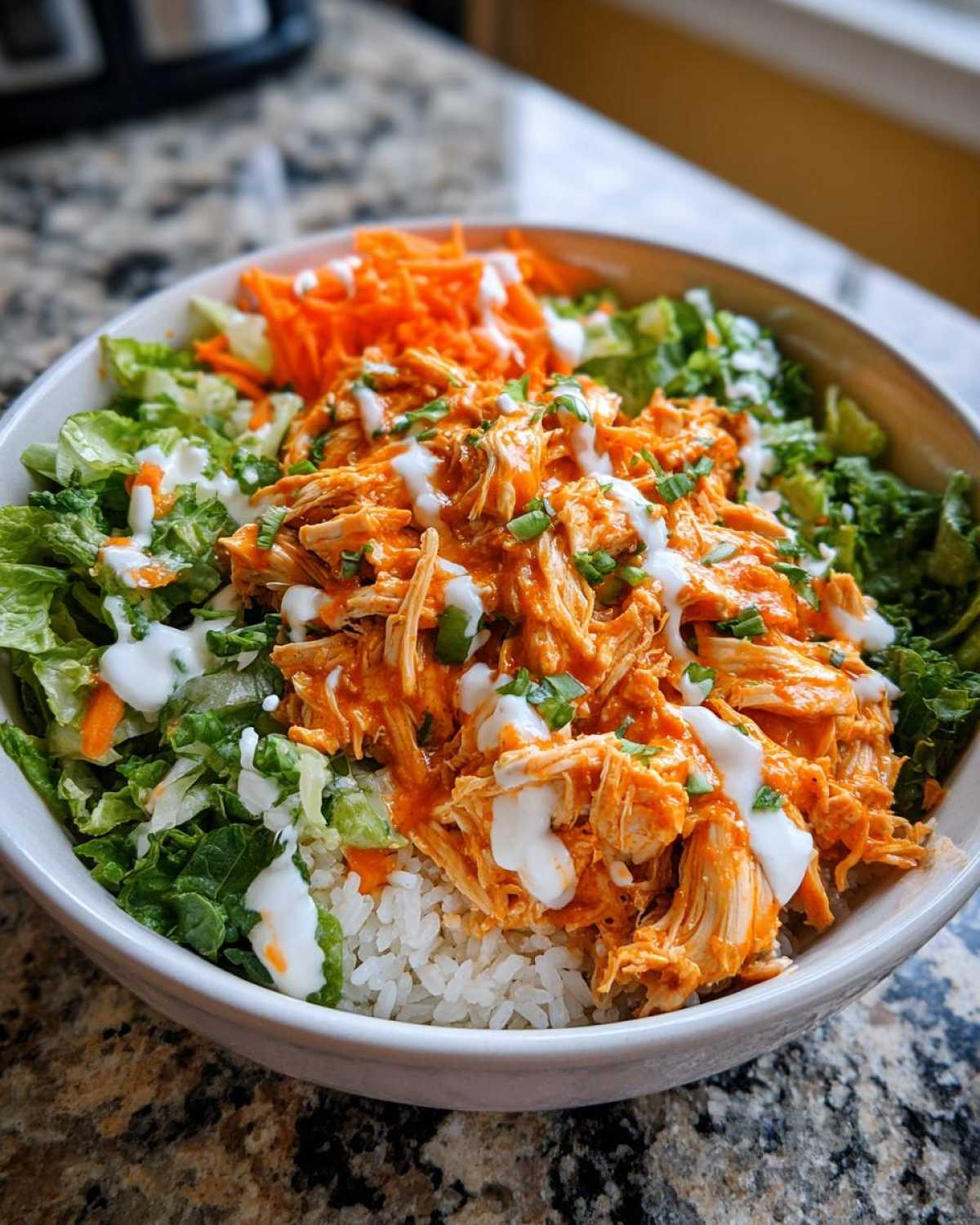 A close-up of a delicious buffalo chicken bowl featuring shredded buffalo chicken over rice, topped with lettuce, carrots, and a ranch drizzle.