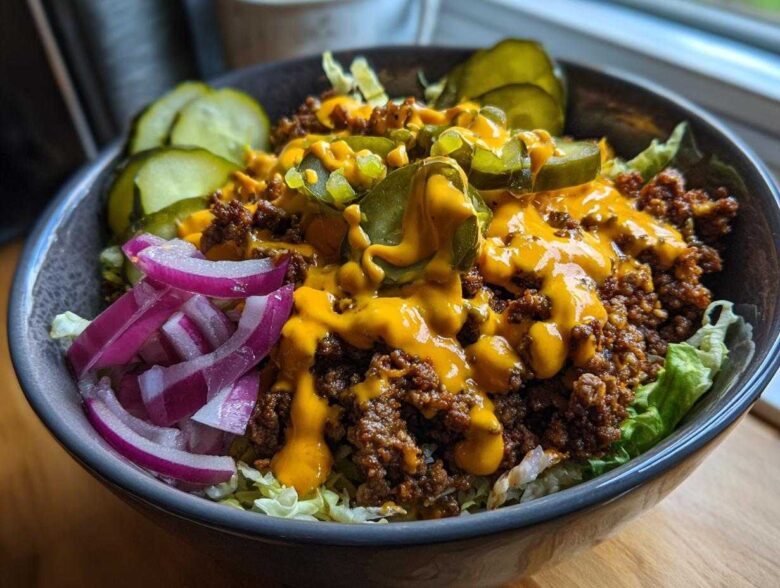 A close-up view of a hearty cheeseburger bowl featuring seasoned ground beef, melted cheese sauce, pickles, and red onion slices over lettuce.