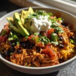 A vibrant, fully loaded chicken burrito bowl featuring shredded chicken, seasoned rice, black beans, avocado slices, salsa, and sour cream.