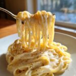 A fork lifting fettuccine noodles generously coated in creamy homemade alfredo sauce and black pepper.