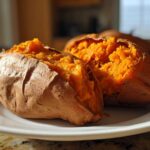 A close-up of a fluffy baked sweet potato split in half, showing its bright orange interior on a white plate.