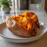 A fluffy baked sweet potato, split open and seasoned with pepper, served on a white plate.