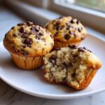 Three fluffy banana chocolate chip muffins on a white plate, one is broken open showing the moist interior.