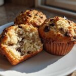Three fluffy banana chocolate chip muffins on a white plate, one broken open to show the moist interior.