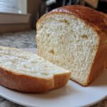 Close-up of a loaf of soft, white condensed milk bread, with one thick slice resting beside it on a white plate.