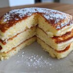 A close-up of a thick stack of fluffy greek yogurt pancakes, cut to show the airy interior, dusted with powdered sugar.