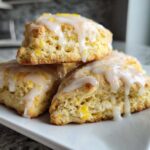 Close-up of fluffy lemon scones stacked on a white plate, drizzled with white lemon glaze and zest.