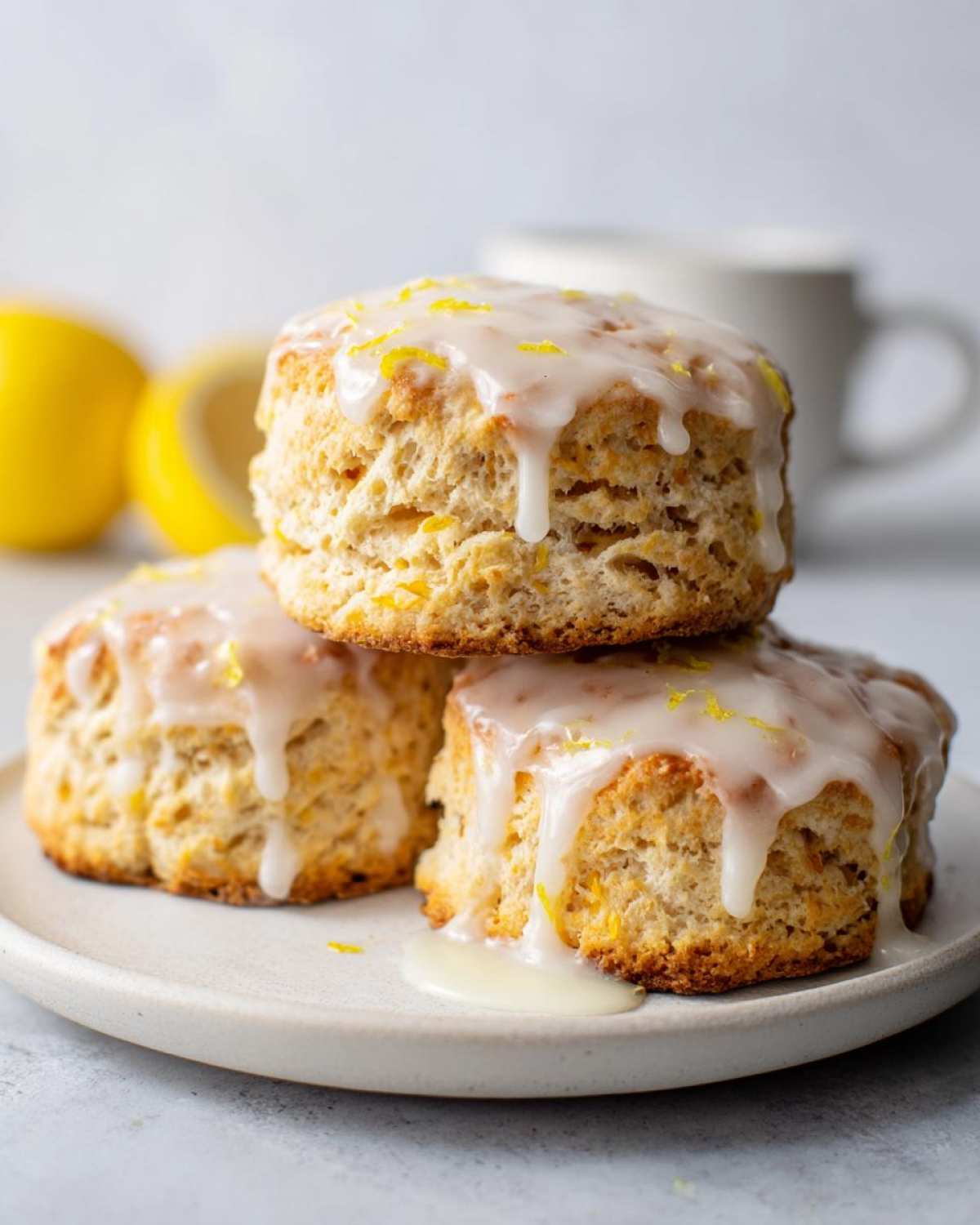 Three fluffy lemon scones stacked on a plate, drizzled with a thick lemon glaze and topped with zest.