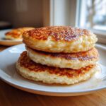 Close-up of three thick, fluffy sourdough pancakes stacked on a white plate, glistening with syrup.