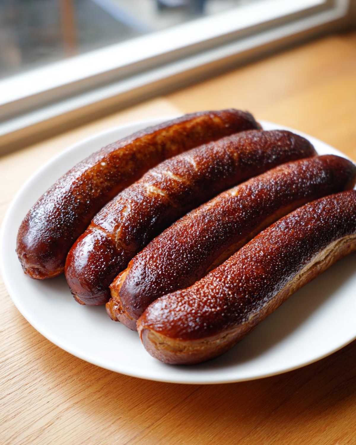 Four freshly baked pretzel dogs with a shiny, dark brown crust arranged on a white plate.