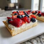 A close-up of a slice of fruit pizza sugar cookie topped with white cream, strawberries, and blueberries.