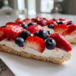 A close-up slice of fruit pizza sugar cookie topped with cream cheese frosting, strawberries, and blueberries.