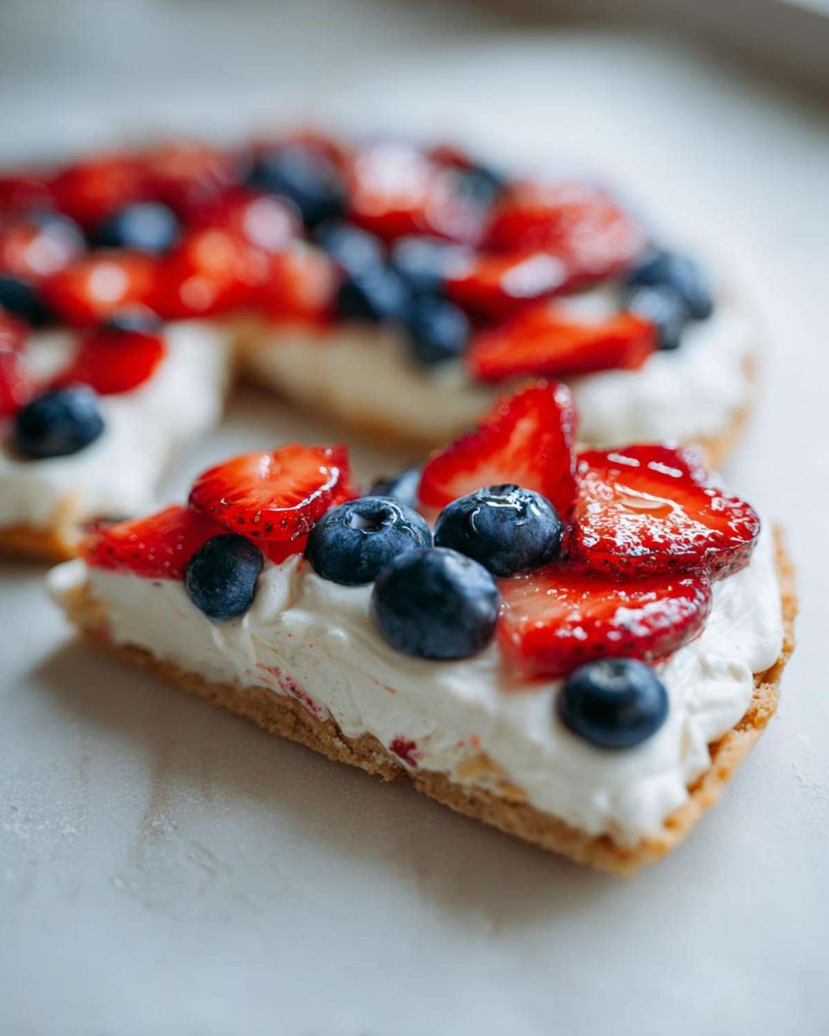 Close-up of a slice of fruit pizza sugar cookie topped with cream cheese frosting, strawberries, and blueberries.