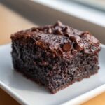 A close-up of a single, dark, fudgy black bean brownies square topped with chocolate chips on a white plate.
