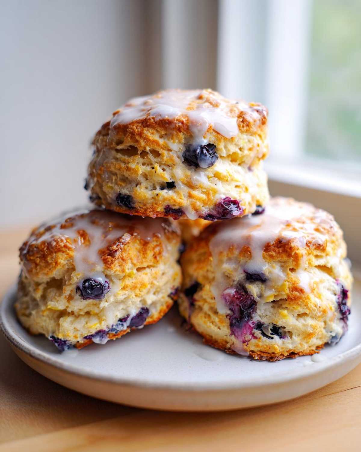 Three fluffy, glazed blueberry scones stacked on a light plate, showcasing visible blueberries inside.