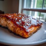 Close-up of a perfectly cooked salmon fillet with a thick, shiny, dark brown glaze, sitting on a white plate.