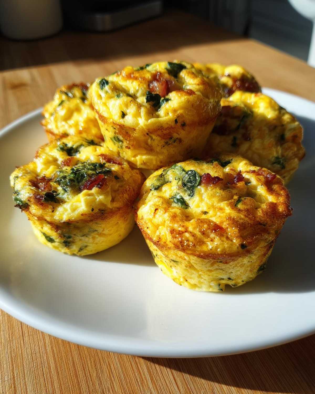 A close-up of several golden brown, baked egg bites with spinach and bacon pieces, resting on a white plate.