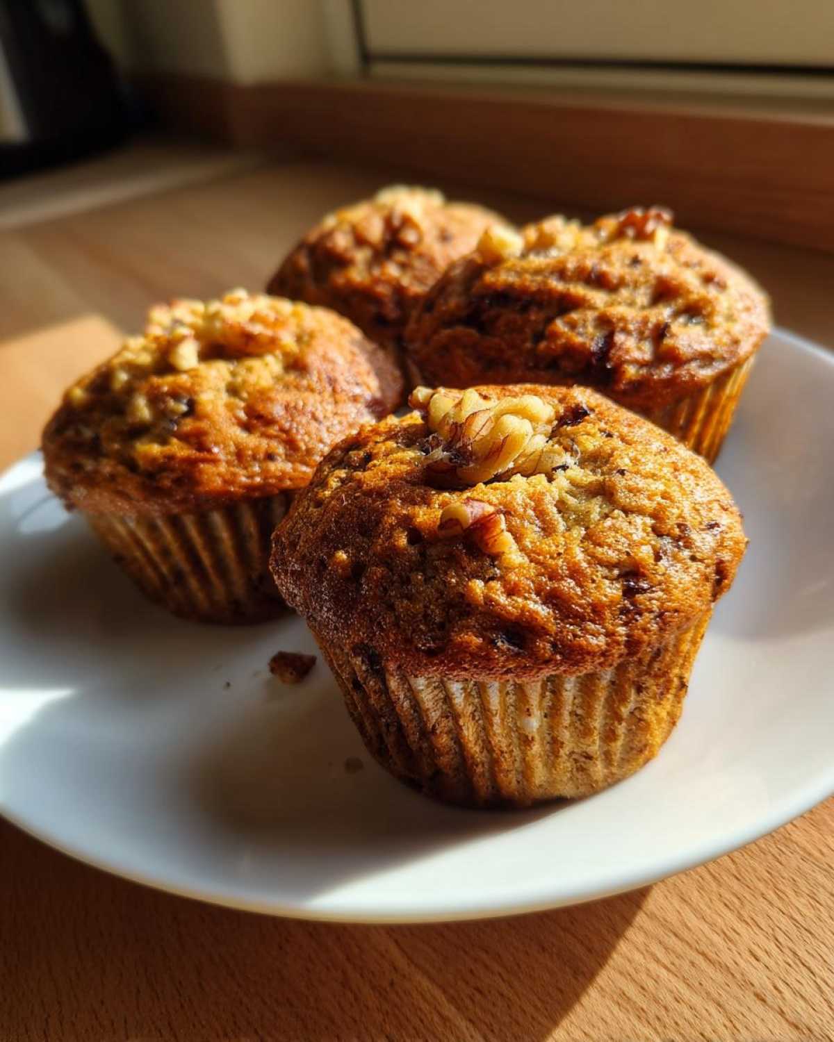 Four freshly baked banana nut muffins topped with walnuts, sitting on a white plate in bright sunlight.