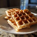Close-up of golden brown high protein waffles stacked on a white plate, catching morning sunlight.