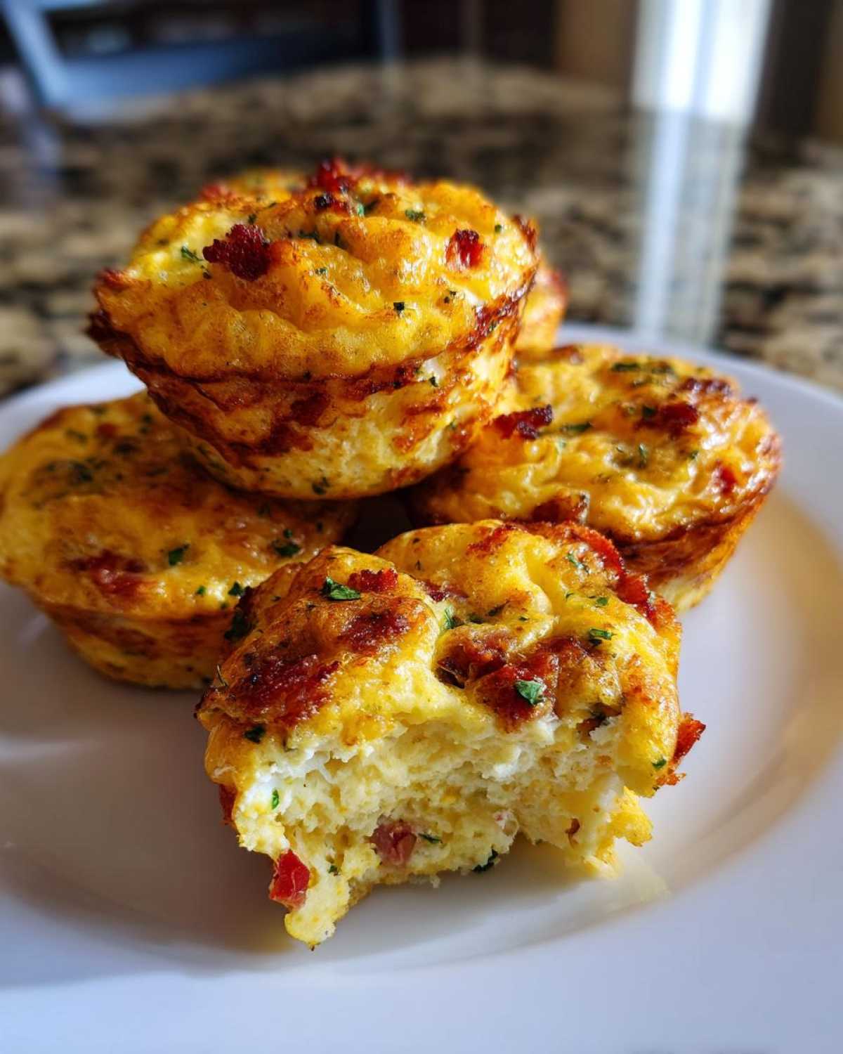 A stack of golden brown muffin tin egg bites, one bitten into, showing fluffy interior with bacon pieces.
