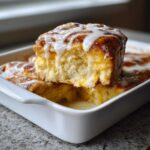 A piece of fluffy Cinnamon roll casserole lifted from a white dish, dripping with sweet white icing.
