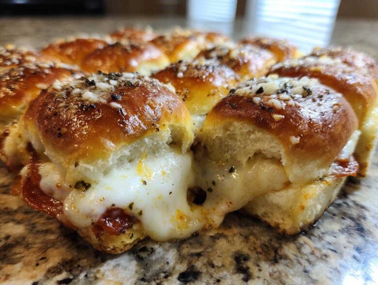 Close-up of gooey pizza sliders showing melted cheese pulling apart between baked rolls, topped with seasoning.