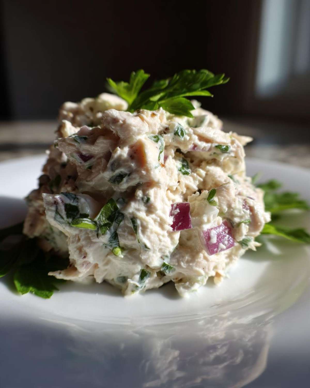 Close-up of a scoop of creamy greek yogurt chicken salad mixed with red onion and parsley on a white plate.