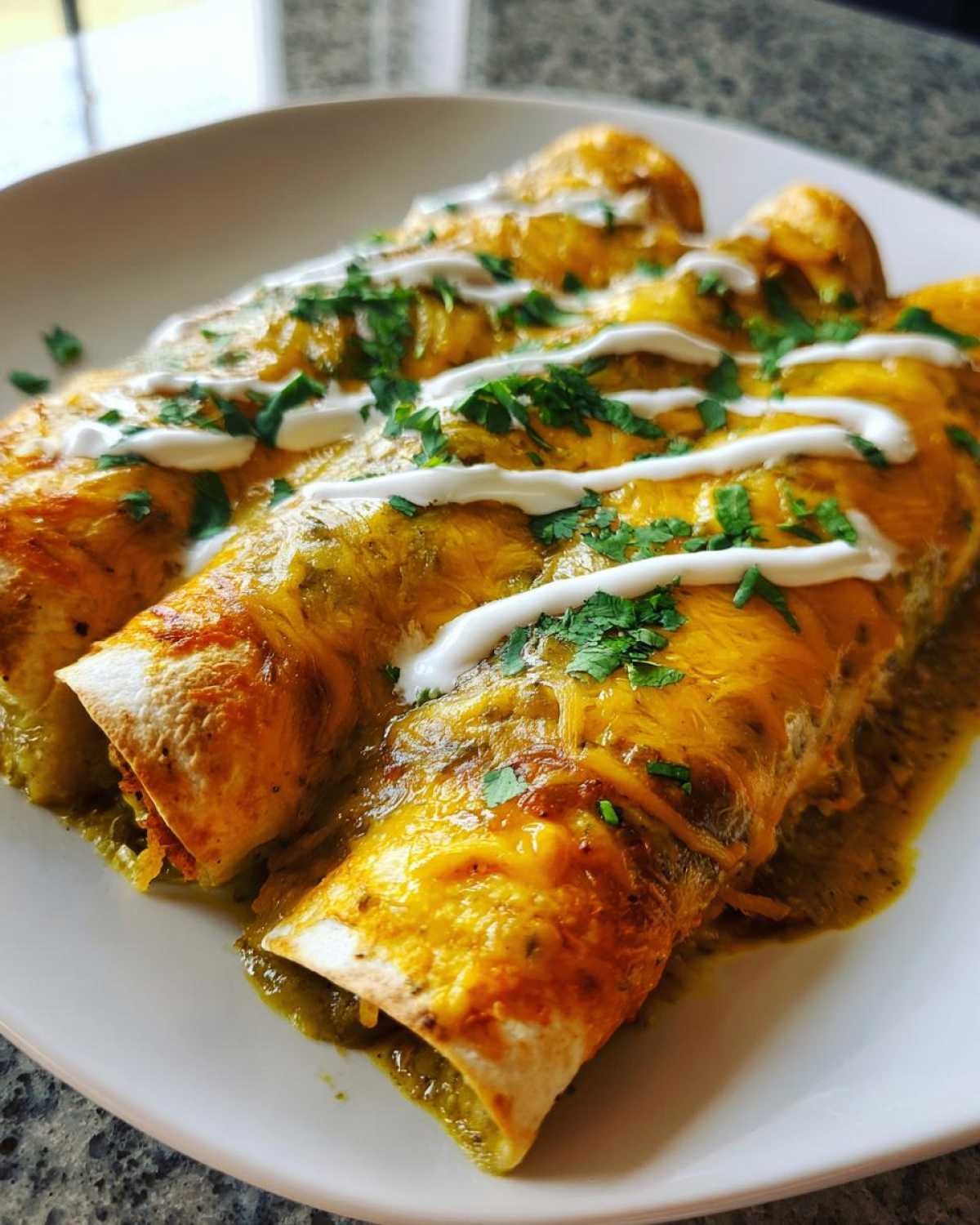 Three baked green chile chicken enchiladas covered in melted cheddar cheese, sour cream drizzle, and cilantro.